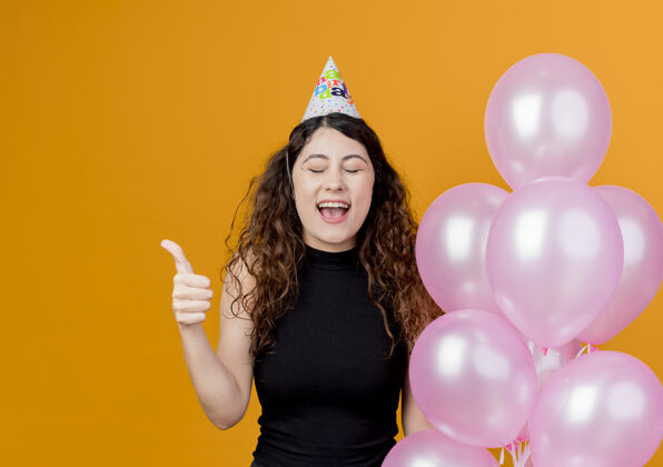 气球年轻漂亮的女人卷发戴着节日帽手持气球疯狂快乐地竖起大拇指站在橙色的墙上生日派对的概念拇指站着疯狂