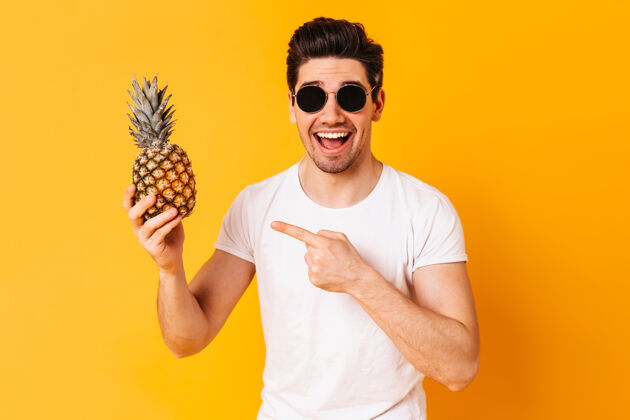 孤独穿着白色t恤和眼镜的快乐男人指着菠萝 在橙色的空间里微笑自信强壮现代