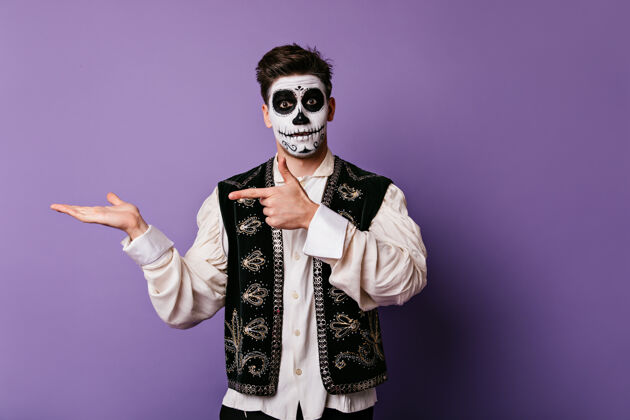 室内一个穿着墨西哥国家队背心的惊讶的家伙用手指指着左边在淡紫色的墙上画着一张画着泰克斯的脸的肖像嘉年华幽灵墨西哥