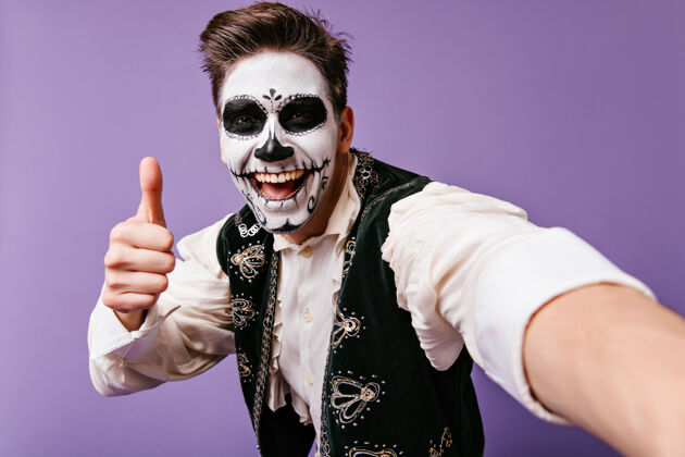 西班牙语在紫色的墙上摆着墨西哥传统人体艺术的快乐的欧洲男人带着僵尸妆的时髦男人在自拍角色节日歌手