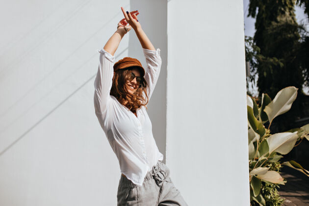 衣服戴着太阳镜 头发卷曲的活跃女人在树环绕的白色建筑旁的街道上跳舞深金发短发放松