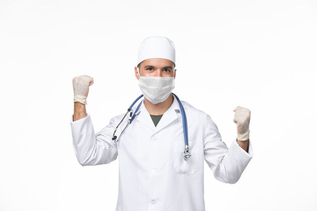 病毒正面图：男医生穿着医疗服 戴着口罩 戴着听诊器 在白墙上欢呼雀跃——疾病大流行病毒男性口罩视图