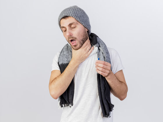 男人咳嗽的年轻病人戴着冬天的帽子和围巾抓住喉咙 拿着隔离在白色的药丸病抱冬天