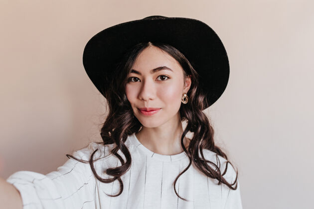 无忧无虑卷曲的中国女人在自拍戴着帽子的亚洲模特在米色背景下摆姿势的前视图年轻时尚中国人
