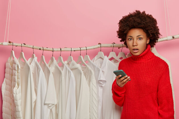 室内尴尬的美国黑人妇女用手机购物 在服装店摆姿势 穿红色毛衣 在衣架旁摆白色衣服黑色震惊一
