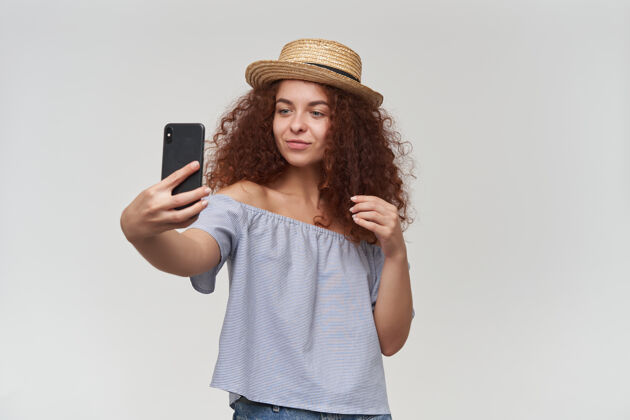 长相漂亮的女人 漂亮的女孩 卷曲的姜黄色头发穿着条纹露肩衬衫和帽子在智能手机上自拍站在白色的墙上女孩手机帽子