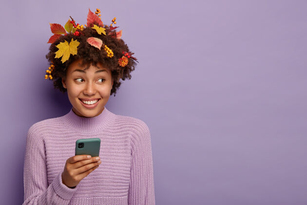 非洲可爱的卷发女人的画像用智能手机 头上有秋叶 神采奕奕 在紫罗兰色的背景上摆姿势叶子电话毛衣