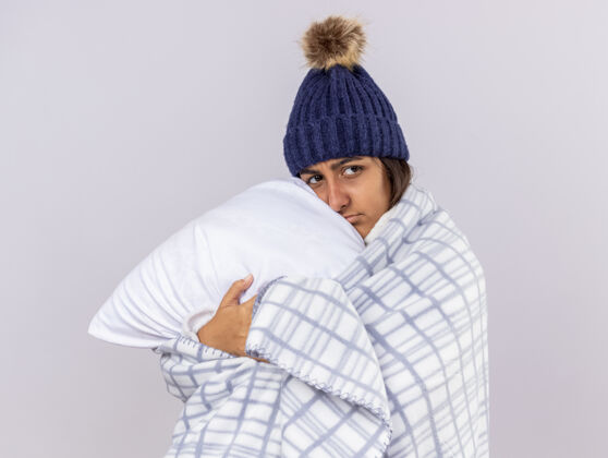 白色悲伤的生病的年轻女孩戴着冬天的帽子 围巾裹着格子呢 抱枕孤零零地躺在白色的床上拥抱穿疾病