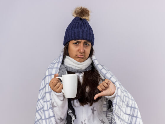 茶未出嫁的生病的年轻女孩戴着冬天的帽子 围巾裹着格子呢 手里拿着一杯茶 拇指朝下拇指年轻杯子