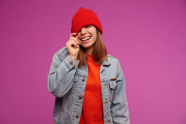 室内有吸引力的 深色头发的成年女孩的肖像穿着红色毛衣 牛仔裤夹克和红色帽子把帽子戴在眼睛上 微笑人们的概念孤立地站在紫色的墙上服装女孩拉