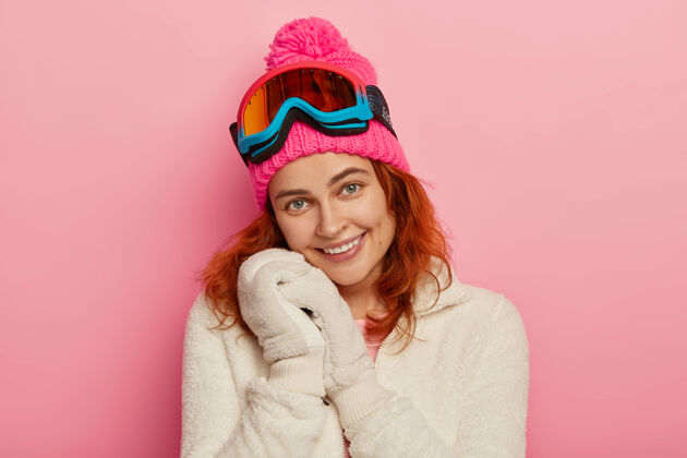 白种人可爱快乐的红发女人的肖像戴着柔软的手套 冬天白色的套头衫 带着绒球和滑雪镜的粉色帽子 温柔的微笑 在粉色背景下与世隔绝手套运动装外套
