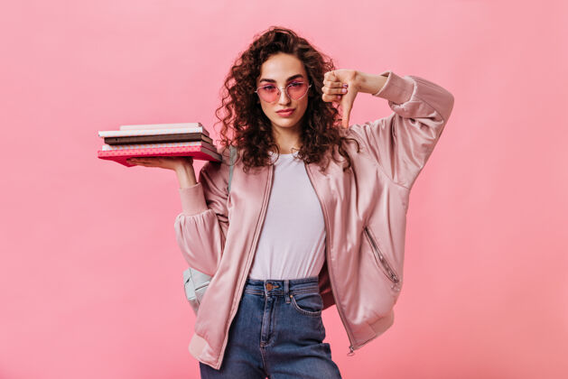 积极戴着粉色太阳镜的女学生拿着书 拇指朝下模特学生笔记本
