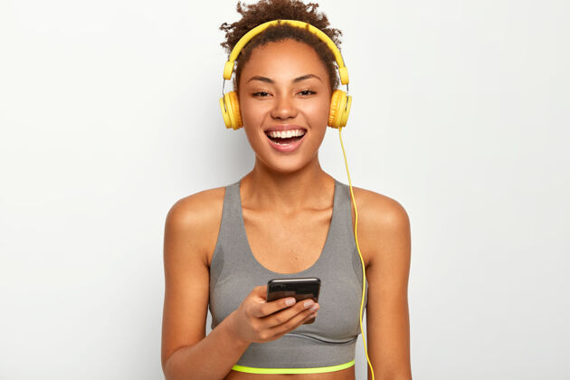 运动穿着运动装的女人 笑容灿烂 使用现代手机 通过耳机听音乐 在白色背景下摆姿势请黑色放松