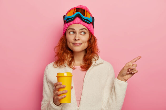 帽子美丽的红发女孩的半身镜头指向右边 向度假地的游客指明方向 有活跃的冬季休息 喝外卖咖啡 戴着滑雪面具 隔离在粉色背景上滑雪板高兴路