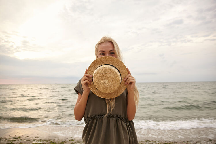 海岸积极的年轻蓝眼睛迷人的白头女性保持她的船帽在自己面前 同时高兴地看着摄像机 在海滩背景隔离提高手姿势
