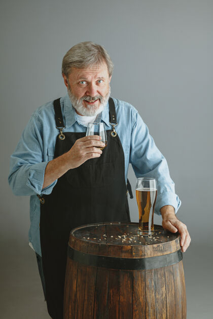 啤酒厂自信的高级酿酒师 在灰色墙壁上的木桶上 用玻璃自制啤酒测试专业酒精
