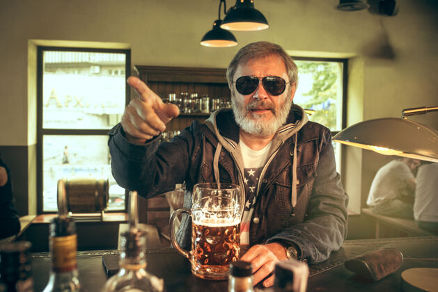 一个大胡子男人在酒吧喝酒 看电视上的体育节目享受我最喜欢的酒和啤酒男人拿着一大杯啤酒坐在桌边胡须男性玻璃