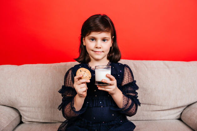 发型小女孩在红墙上喝牛奶孩子吃饼干的室内镜头饥饿微笑情绪