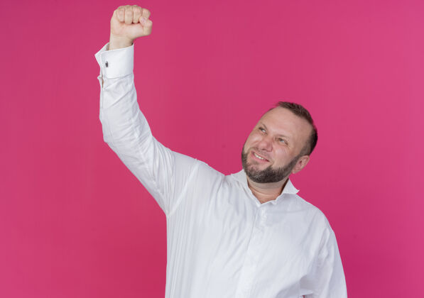 兴奋一个留着胡子的男人穿着白衬衫 握紧拳头 高兴而兴奋地站在粉红色的墙上为自己的成功而欢呼男人成功欢喜
