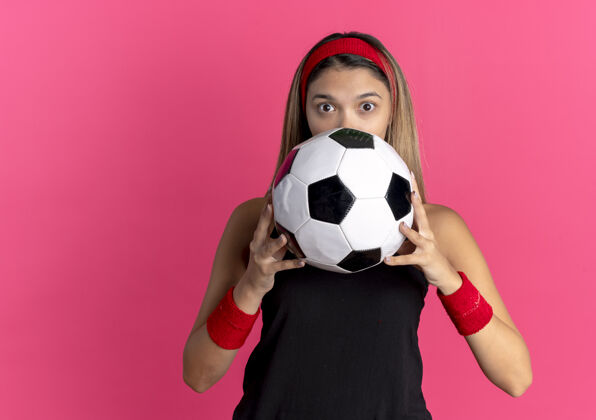 年轻身穿黑色运动服 头戴红色头带 手持足球的年轻健身女孩站在粉色的墙上 脸上藏着惊讶的表情健身看足球