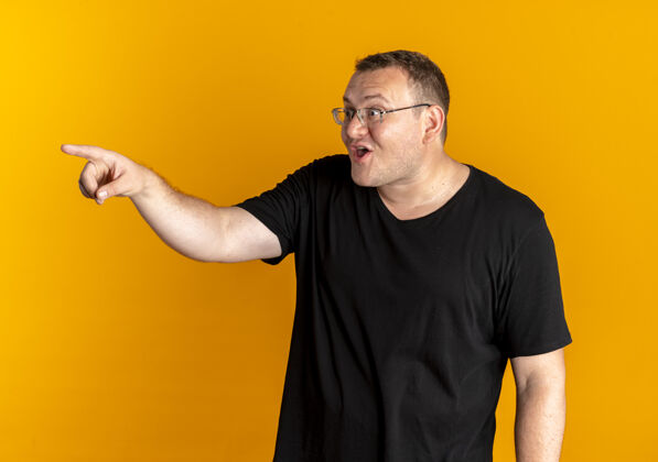 兴奋一个戴着眼镜 身穿黑色t恤的超重男子用手指指着站在橙色墙上的某个快乐而兴奋的东西男人手指穿什么