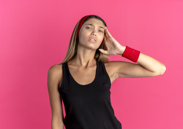 站身穿黑色运动服 头戴红色头巾的年轻健身女孩站在粉红色的墙上 看上去很不舒服 头痛年轻看健康