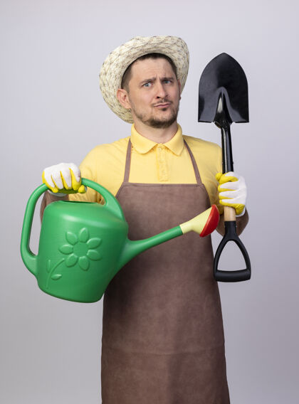 浇水年轻的园丁 穿着连体衣 戴着帽子 戴着工作手套 手里拿着水罐和铲子 感到困惑手套帽子花园