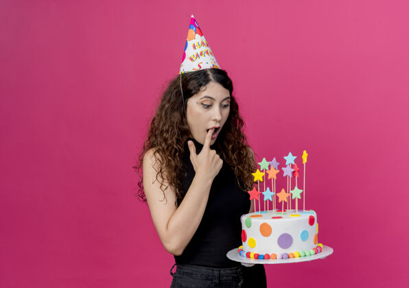 惊喜一个年轻漂亮的女人 卷发 戴着节日帽 拿着生日蛋糕 看着它 站在粉色的墙上 惊讶地看着生日派对的概念帽子惊奇抱着