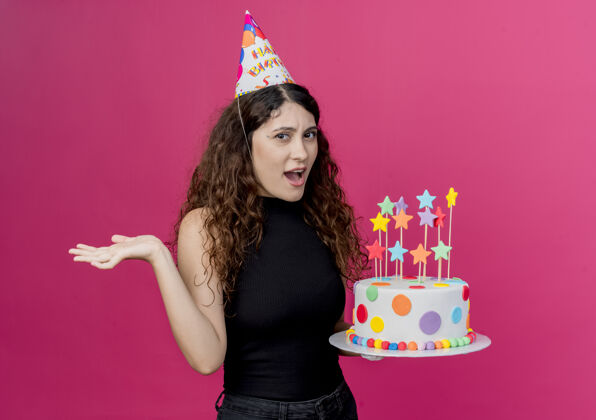 帽子一个年轻漂亮的女人 卷发 戴着节日帽 手里拿着生日蛋糕 站在粉色的墙上 困惑着生日派对的概念年轻卷发困惑