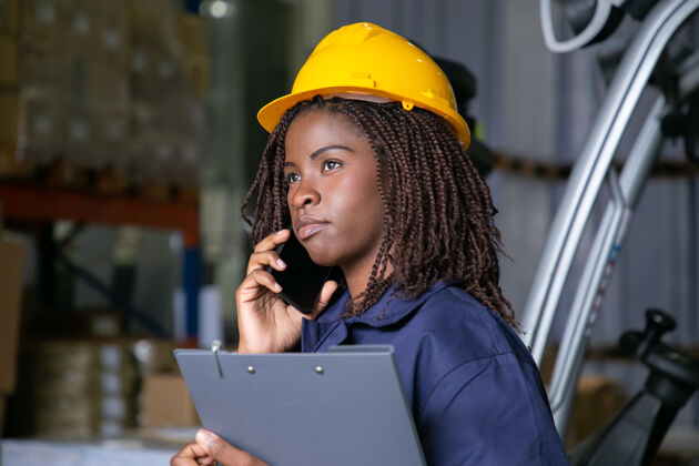 多样性戴着安全帽的沉思的黑人女工程师站在仓库里 讲着手机背景是货物的架子复制空间劳动或通讯概念安全帽工程师谈话