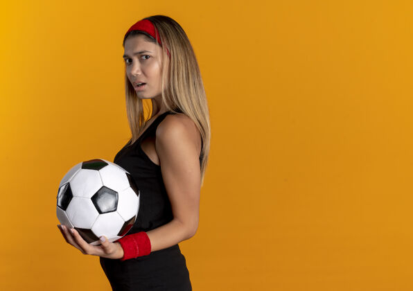 抱身穿黑色运动服 头戴红色头巾 手持足球的年轻健身女孩站在橘色的墙上不高兴地站着女孩年轻运动装