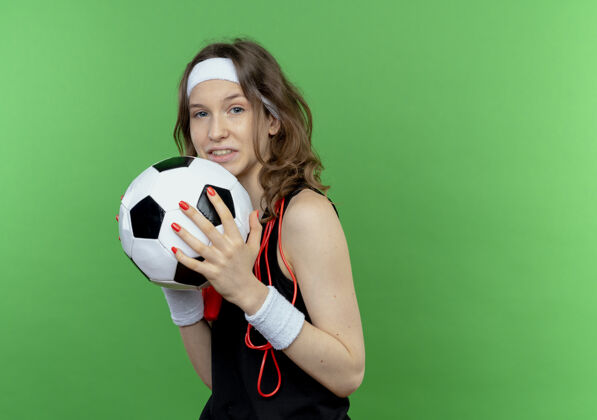 站着身穿黑色运动服的年轻健身女孩 头上系着头带 脖子上系着跳绳 手里拿着足球 微笑着站在绿色的墙上健身女孩球