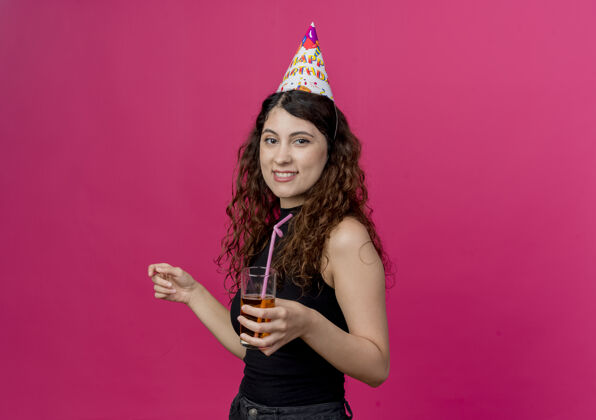 卷发一位年轻漂亮的女士 一头卷发 戴着节日帽 手里拿着鸡尾酒 微笑着 站在粉色的墙上微笑年轻帽子