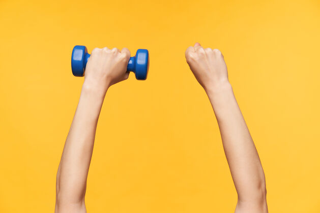 女性皮肤白皙强壮的女性举起双手 在黄色背景下摆姿势 用蓝色哑铃锻炼肌肉的水平照片身体护理配件特写