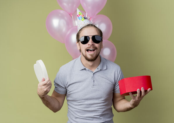 举行戴着节日帽的年轻人举着礼盒庆祝生日快乐而兴奋地微笑着 手里拿着气球站在轻墙上气球光年轻