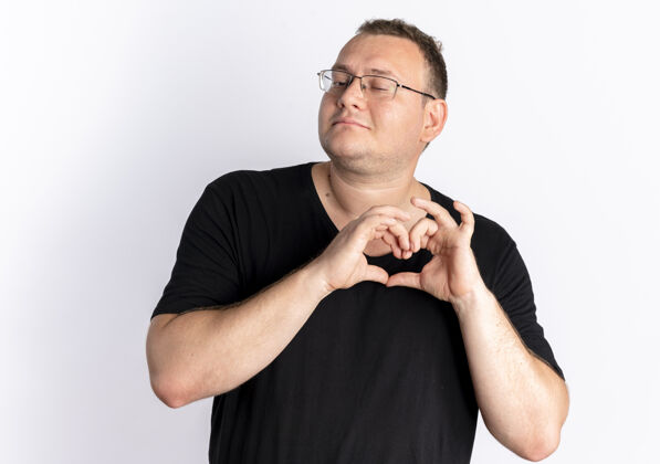 超重戴眼镜的超重男子身穿黑色t恤 站在白墙上做心脏手势 用手指感受积极的情绪眼镜心做