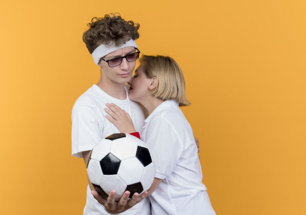 站着年轻的运动夫妇伤心的女人拥抱男子 而他拿着足球站在橙色的墙上拥抱球悲伤