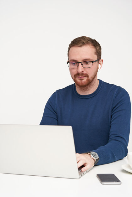玻璃沉思的年轻漂亮的胡子 金发 戴着眼镜 正坐在白色背景上 在键盘上打字 专心工作水笔记本电脑毛衣