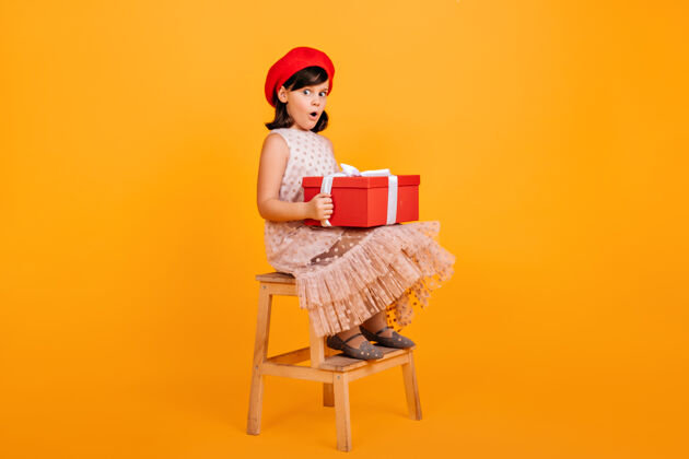 惊喜穿着裙子的漂亮小女孩坐在椅子上 手里拿着一个大礼盒有生日礼物的法国小孩黄色隔离庆祝椅子