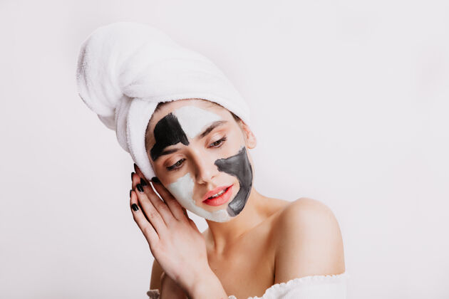 毛巾戴着泥塑面具的冷静女士在白墙上摆姿势做晚间卫生的女人的画像头发眉毛护理