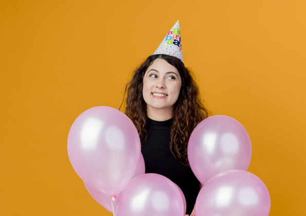站着一位年轻漂亮的女士 卷发 戴着节日帽 手持气球 站在橙色的墙上 快乐而兴奋地庆祝生日帽子女人空气