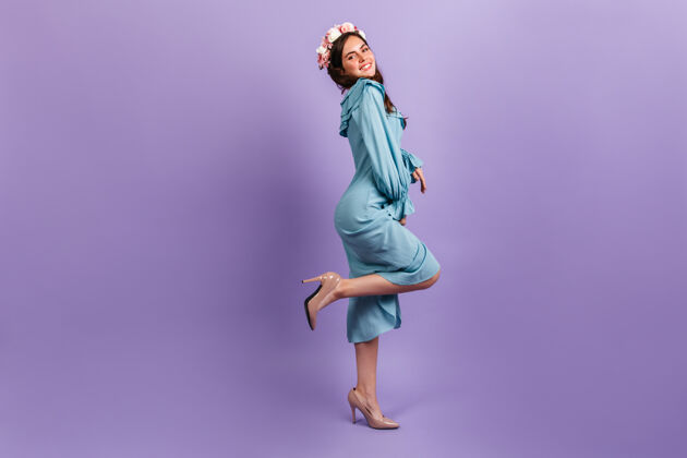 服装穿着高跟鞋和迷笛裙的年轻黑发女郎的全长照片头发上插着鲜花的女模特在淡紫色的墙上微笑魅力肖像模特
