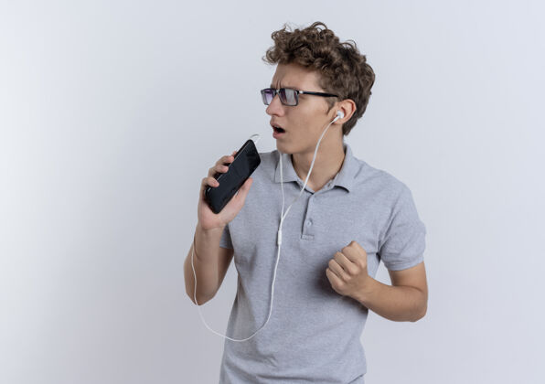 麦克风穿着灰色马球衫的年轻人戴着耳机拿着智能手机当麦克风站在白墙上唱歌马球年轻歌手