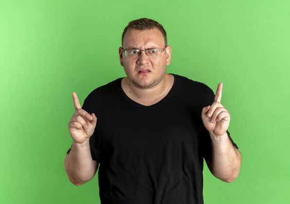 手指戴眼镜的超重男子戴着黑色t型眼镜 看起来很困惑 手指站在绿色的墙上戴着指指点点男人