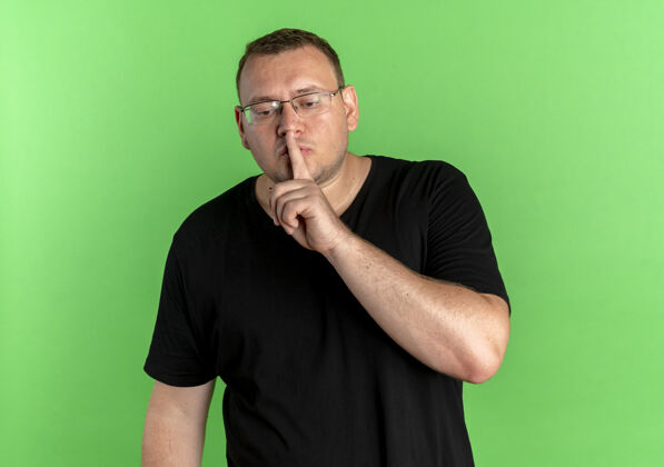男人戴眼镜的超重男子身穿黑色t恤 手指放在嘴唇上 站在绿色的墙上做着沉默的手势超重制造站立
