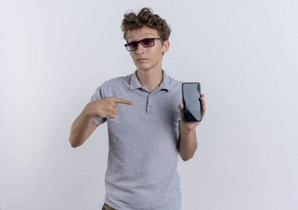 手指一个穿着灰色马球衫的年轻人站在白色的墙上 用食指指着智能手机 感到困惑移动马球站