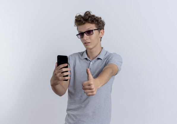 拿着穿着灰色马球衫的年轻人举着智能手机 竖起大拇指站在白色的墙上马球年轻的站着
