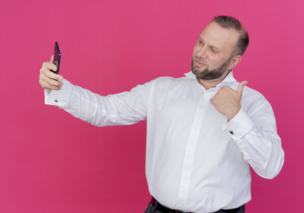 穿一个留着胡子的男人穿着白衬衫站在粉色的墙上自拍 竖起大拇指自拍胡子秀