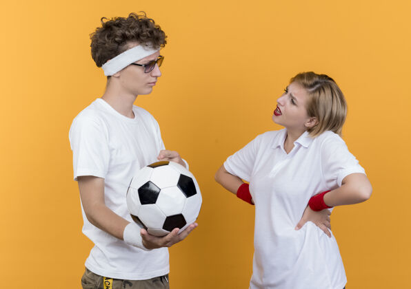 站年轻的运动型情侣严肃的男人拿着足球看着站在橘色墙壁上不高兴的女友严重情侣女朋友