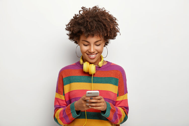 年轻美丽的非洲裔美国女人的照片集中在智能手机设备中 喜欢在线聊天 下载音乐到播放列表中 戴着耳机收听 卷曲的黑发 穿着休闲装姿势高兴正面